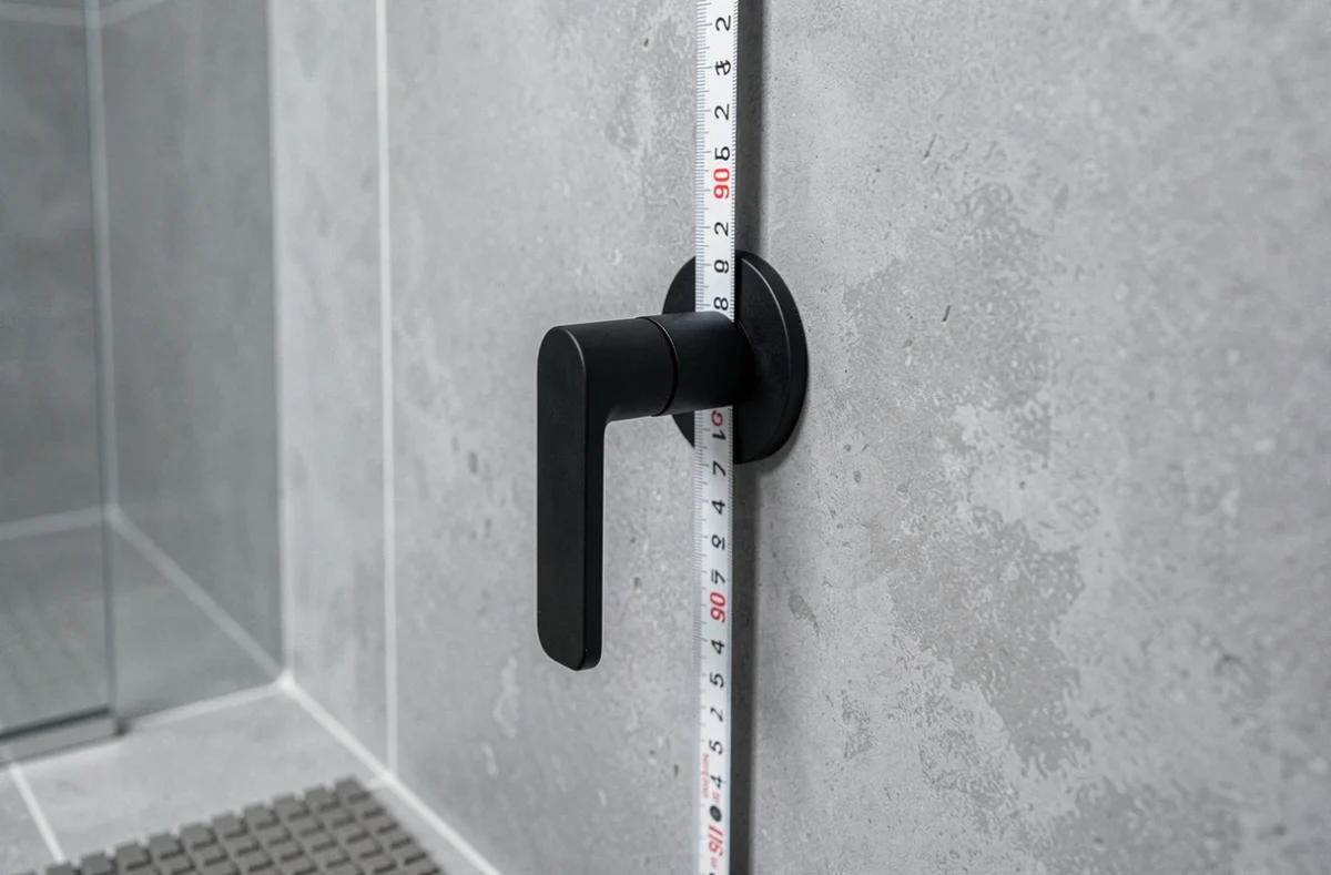 Gros plan sur un mitigeur douche en noir mat installé à la hauteur réglementaire PMR de 90 cm avec mètre ruban sur carrelage gris